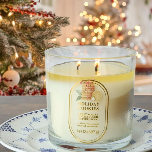 A lit \“Holiday Cookies\“ scented candle featuring sweet vanilla, cookie dough, and buttercream aromas is placed on a blue and white plate, with a blurred Christmas tree in the background.