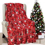 A red holiday-themed blanket with festive designs like Santa, polar bears, and \“Ho Ho Ho\“ text is draped over a chair next to a decorated Christmas tree with ornaments and presents underneath.