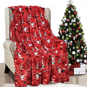 A red holiday-themed blanket with festive designs like Santa, polar bears, and \“Ho Ho Ho\“ text is draped over a chair next to a decorated Christmas tree with ornaments and presents underneath.