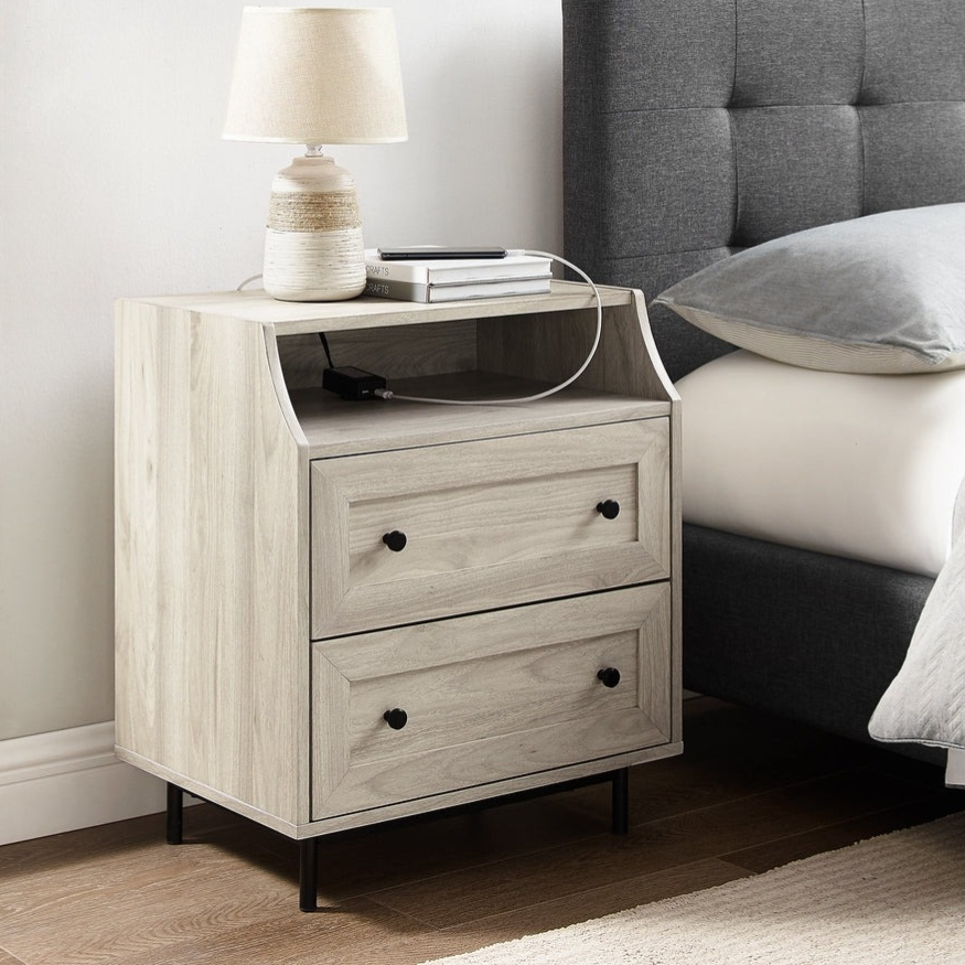 A light-colored wooden nightstand with two drawers and an open shelf, featuring a table lamp on top. It's positioned next to a gray upholstered bed with a quilted headboard.