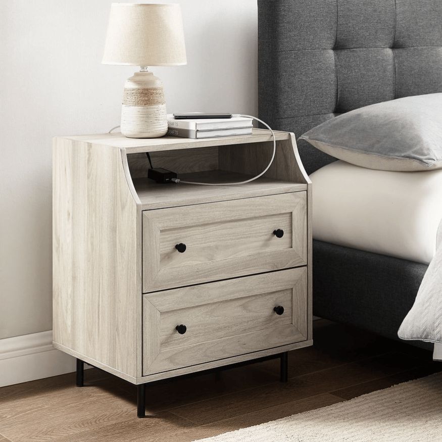 A light-colored wooden nightstand with two drawers and an open shelf, featuring a table lamp on top. It's positioned next to a gray upholstered bed with a quilted headboard.