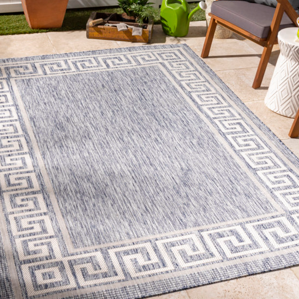 8' x 10' outdoor geometric rug with a blue and white design, featuring a Greek key border pattern, set on a patio with chairs and plants.