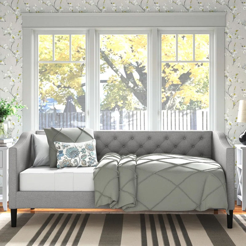 A gray upholstered twin daybed features a tufted back and arms, accented with plush cushions and a patterned throw pillow, against floral wallpaper and large windows with a nature view.