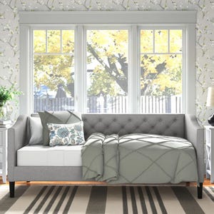A gray upholstered twin daybed features a tufted back and arms, accented with plush cushions and a patterned throw pillow, against floral wallpaper and large windows with a nature view.