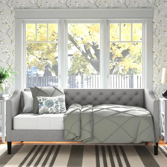 A gray upholstered twin daybed features a tufted back and arms, accented with plush cushions and a patterned throw pillow, against floral wallpaper and large windows with a nature view.
