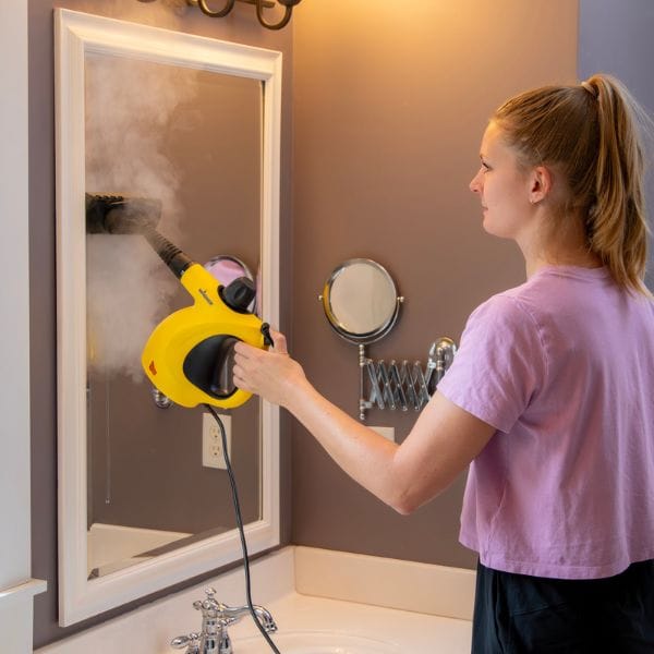 A woman uses a Wagner Handheld Steamer with a yellow body to clean a mirror, emitting steam. The steamer is compact and features an ergonomic handle for convenient use in household cleaning tasks.