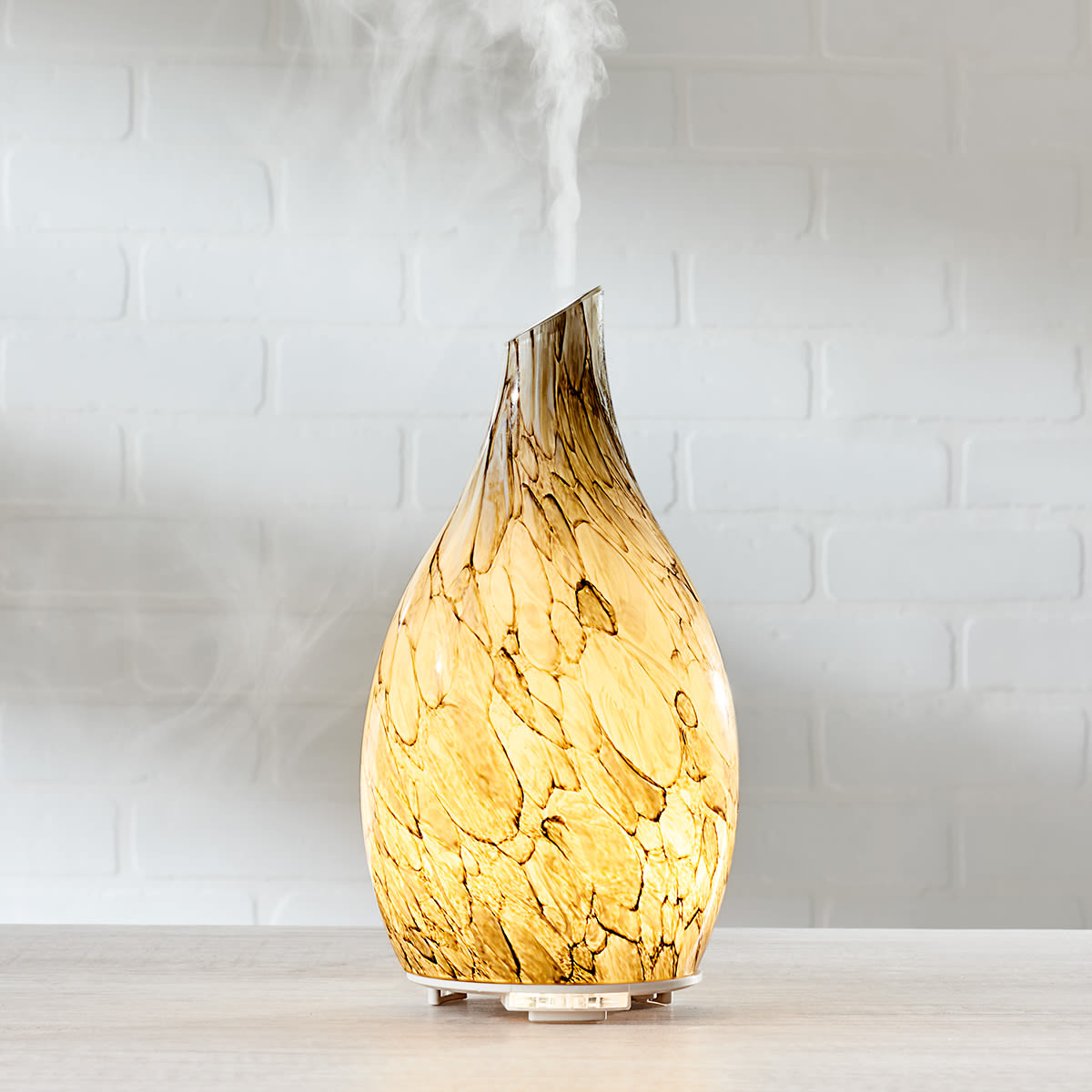 A sand-blasted glass diffuser with a marbled pattern emits mist from a narrow, angled top, creating an illuminated, ambient glow. It's elegantly rounded and sits on a flat base.
