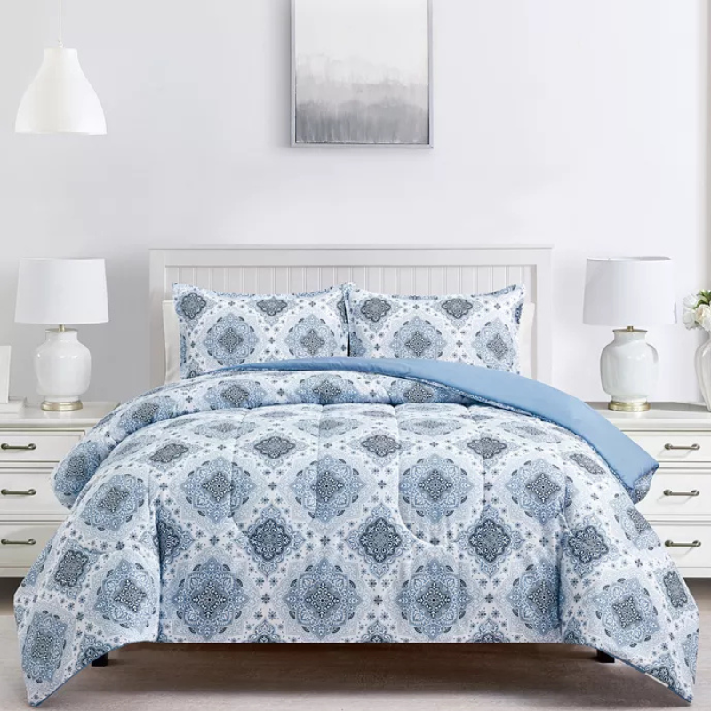 A bed is styled with a blue and white comforter set featuring a geometric pattern. The room includes two white lamps with gold accents, a white headboard, and a gray decorative wall piece above the bed.