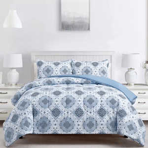 A bed is styled with a blue and white comforter set featuring a geometric pattern. The room includes two white lamps with gold accents, a white headboard, and a gray decorative wall piece above the bed.