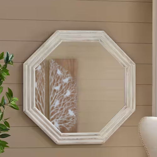 Octagonal white wooden-framed wall mirror, set against a beige shiplap wall, with a glimpse of a leafy artwork in the reflection.