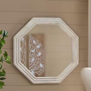 Octagonal white wooden-framed wall mirror, set against a beige shiplap wall, with a glimpse of a leafy artwork in the reflection.