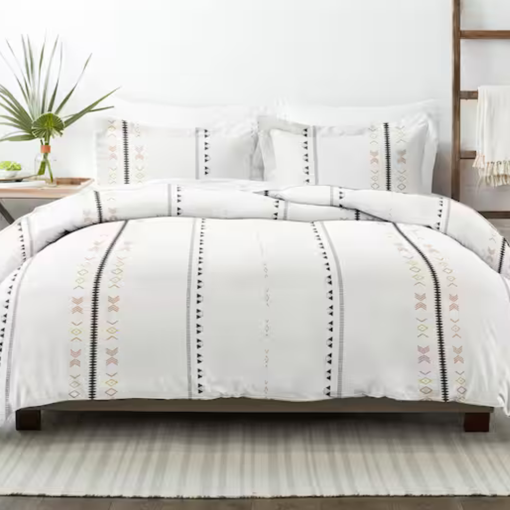 A bed features a white comforter and pillows with vertical geometric patterns in black and soft colors. A bedside table holds a vase with greenery, and there is a wooden ladder with a draped blanket nearby, all atop a striped rug.