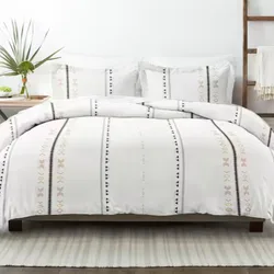 A bed features a white comforter and pillows with vertical geometric patterns in black and soft colors. A bedside table holds a vase with greenery, and there is a wooden ladder with a draped blanket nearby, all atop a striped rug.