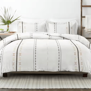 A bed features a white comforter and pillows with vertical geometric patterns in black and soft colors. A bedside table holds a vase with greenery, and there is a wooden ladder with a draped blanket nearby, all atop a striped rug.