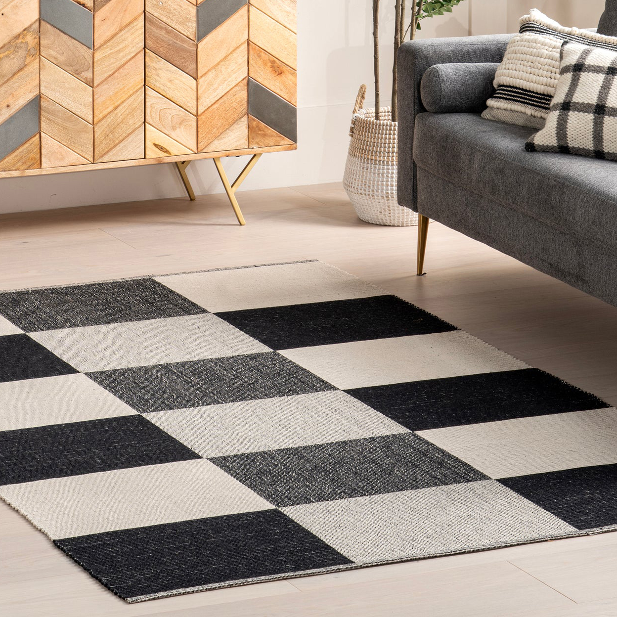 A checkered area rug with a black and cream design is placed near a gray sofa with patterned pillows, set in a room featuring a wooden cabinet with a chevron pattern and decorative elements.