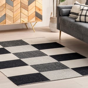 A checkered area rug with a black and cream design is placed near a gray sofa with patterned pillows, set in a room featuring a wooden cabinet with a chevron pattern and decorative elements.