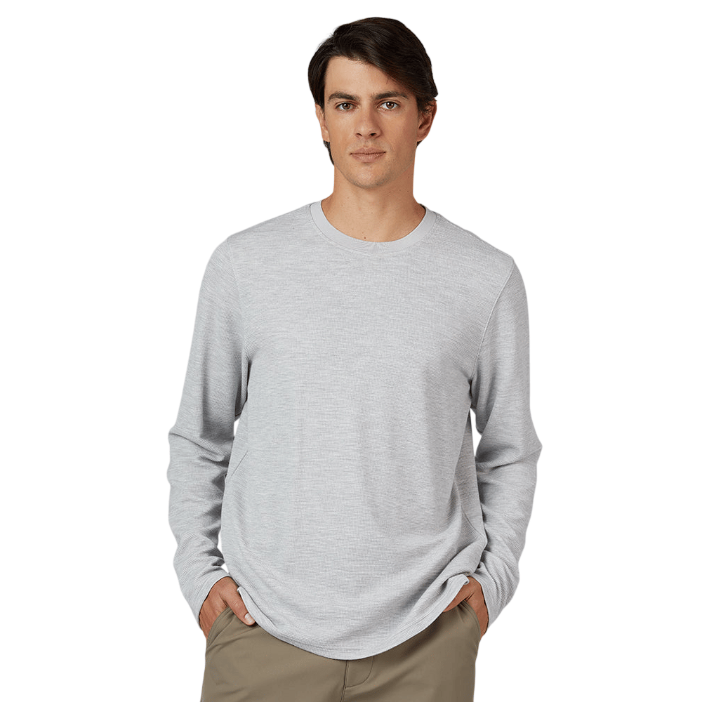 A man is wearing a light gray long-sleeve crew neck shirt and khaki pants, with hands in pockets, set against a plain gray background.