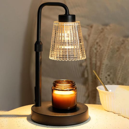A black lamp with a glass shade is shown warming a lit candle in a brown jar on a round base, next to a white bowl with a spoon.