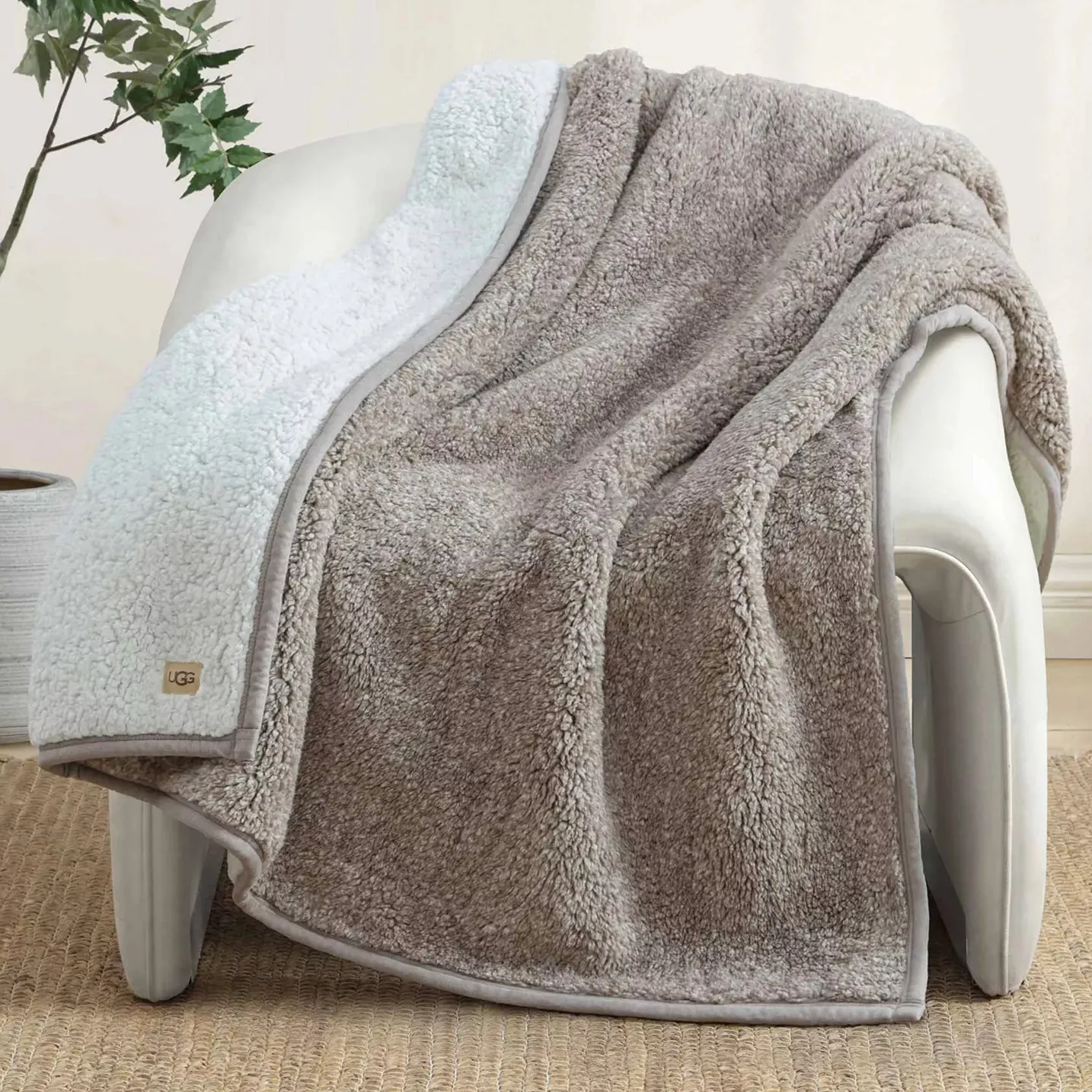 A cozy, dual-textured, tan and white throw blanket with a small UGG logo is draped over a modern, white chair on a woven rug, next to a potted plant.