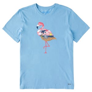 Light blue T-shirt featuring a graphic of a pink flamingo with a sunset beach scene, including palm trees, a chair, and an ocean, integrated into the flamingo's body. Text reads \“Life is Good.\“