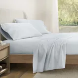 A bed with light blue sheets, two matching pillows, and a wooden bedside table with books and a small bowl. The room has a large window with a view of greenery.