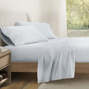 A bed with light blue sheets, two matching pillows, and a wooden bedside table with books and a small bowl. The room has a large window with a view of greenery.