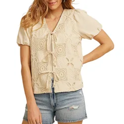 A woman is wearing a cream-colored crochet vest with tie closures over a short-sleeved blouse, paired with light blue distressed denim shorts.