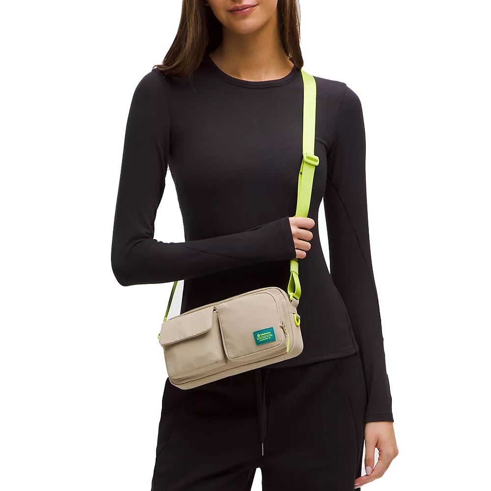 A woman wears a black long-sleeve top and black pants, carrying a beige crossbody bag with a neon green strap and a small front pocket with a logo patch.