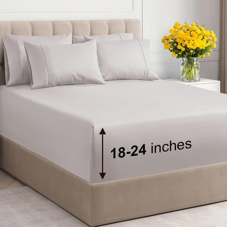 Bed with light gray sheets and four pillows, featuring a fit for mattresses 18-24 inches deep. A vase with yellow flowers is on the nightstand.