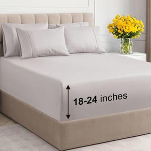 Bed with light gray sheets and four pillows, featuring a fit for mattresses 18-24 inches deep. A vase with yellow flowers is on the nightstand.