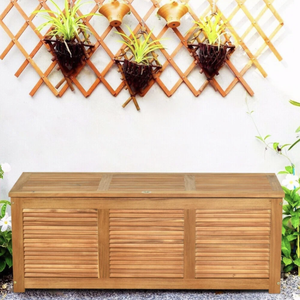 Outdoor storage bench with a slatted design, placed against a white wall with a decorative trellis featuring hanging plants and copper-colored pots.