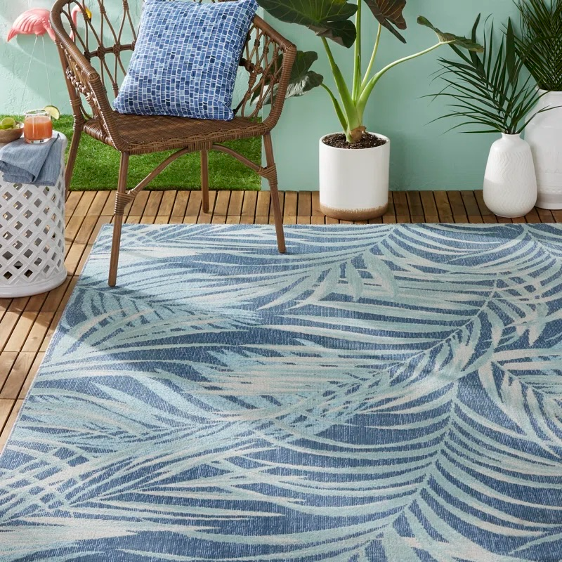A wicker chair with a blue patterned cushion sits on a wooden deck, accompanied by a blue and white palm leaf-patterned rug. Nearby are two white vases and a potted plant.