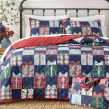 A bed features a patchwork quilt and matching shams with a gift box pattern in red, blue, and green tones, complemented by a red quilted lining. A matching tote with handles is placed on the bed.