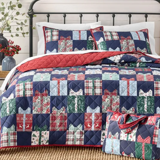 A bed features a patchwork quilt and matching shams with a gift box pattern in red, blue, and green tones, complemented by a red quilted lining. A matching tote with handles is placed on the bed.