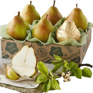 A set of six Harry & David pears, one wrapped in gold foil, displayed in a branded box with green paper. The image also includes sliced pear pieces and leafy branches for decoration.