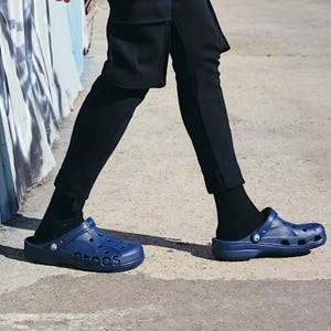 A person is wearing black Crocs with a strap and black socks, paired with black trousers.