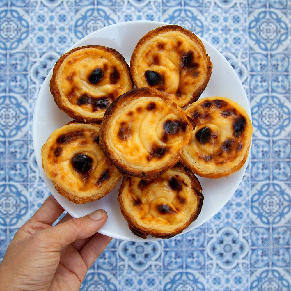 A plate of traditional Portuguese pastéis de nata, creamy custard tarts with golden, flaky pastry, is held against a background of blue and white patterned tiles.