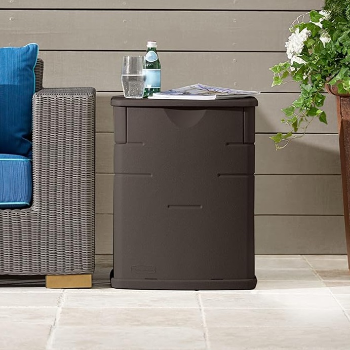 A brown plastic storage unit with a flat top holding a glass of water, bottle, and a newspaper, beside a wicker chair with blue cushions.