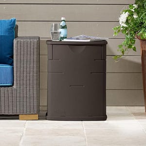 A brown plastic storage unit with a flat top holding a glass of water, bottle, and a newspaper, beside a wicker chair with blue cushions.