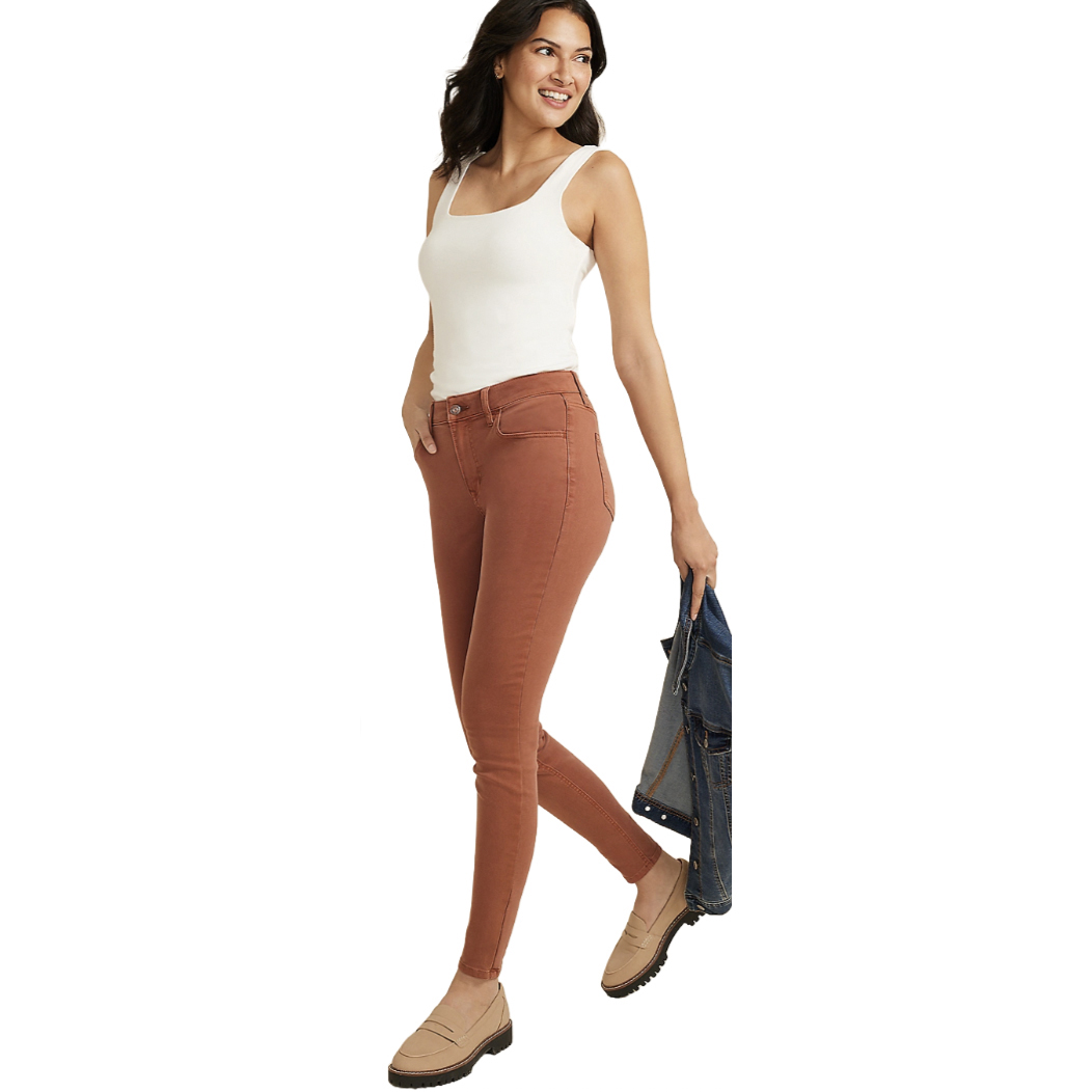 A woman wears a white tank top, rust-colored skinny jeans, and beige loafers, holding a denim jacket.