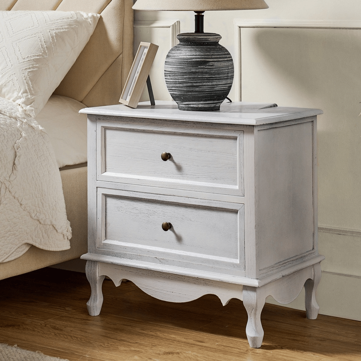A rustic white bedside table with two drawers and round knobs is accompanied by a textured table lamp, situated next to a beige upholstered bed.