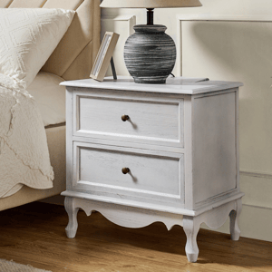 A rustic white bedside table with two drawers and round knobs is accompanied by a textured table lamp, situated next to a beige upholstered bed.