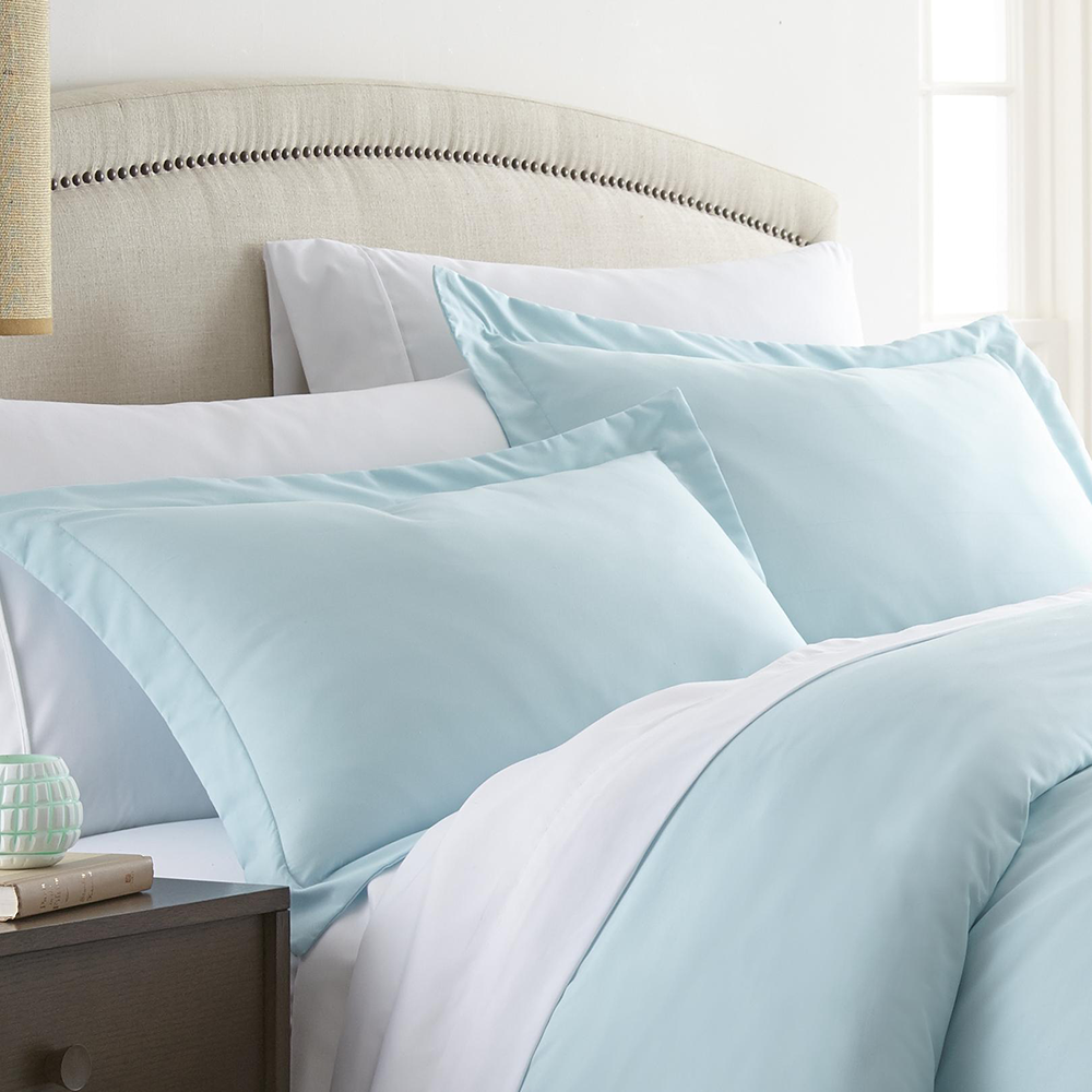 Two light blue pillow shams with a satin-like texture, featuring a simple stitched border. They are placed on a bed with a beige upholstered headboard and are set against crisp white sheets.