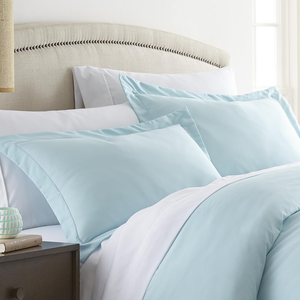 Two light blue pillow shams with a satin-like texture, featuring a simple stitched border. They are placed on a bed with a beige upholstered headboard and are set against crisp white sheets.