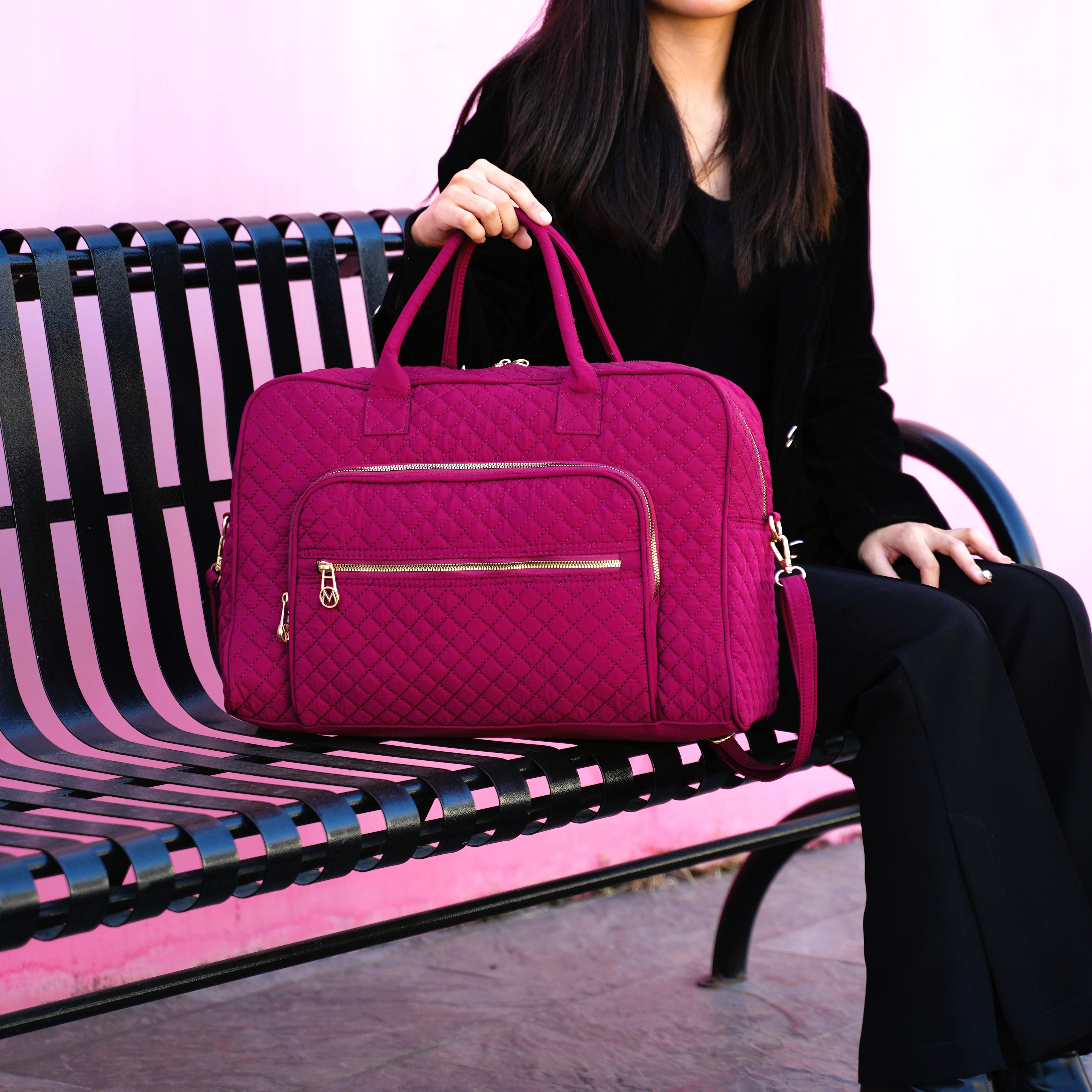 A woman is seated on a bench holding a magenta quilted handbag with multiple zippered compartments and handles.