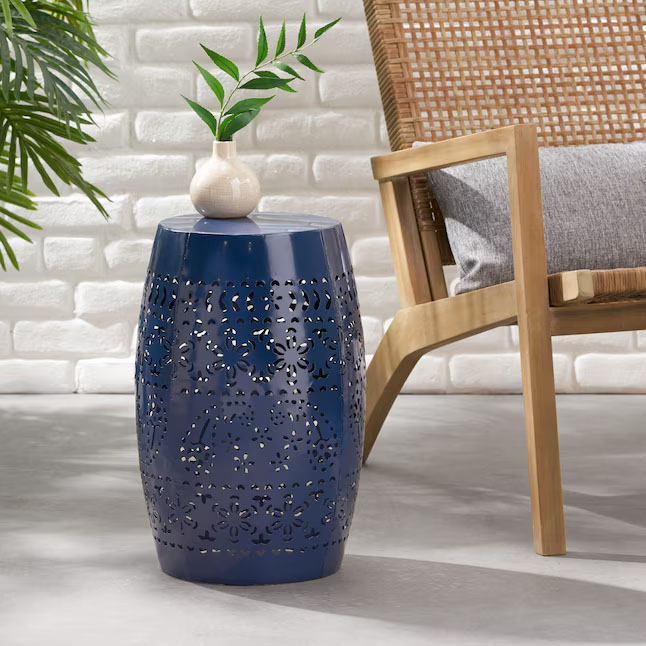A blue ceramic garden stool features intricate cut-out patterns on its body. It is topped with a small white vase holding green foliage, next to a wooden chair with a woven design and gray cushion, set against a white brick wall.