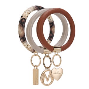 Three round keychain wristlets are shown, each with a different design: brown leather, beige faux leather, and snakeskin pattern. They feature gold accents and charms with the text \“MIA K. COLLECTION.\“