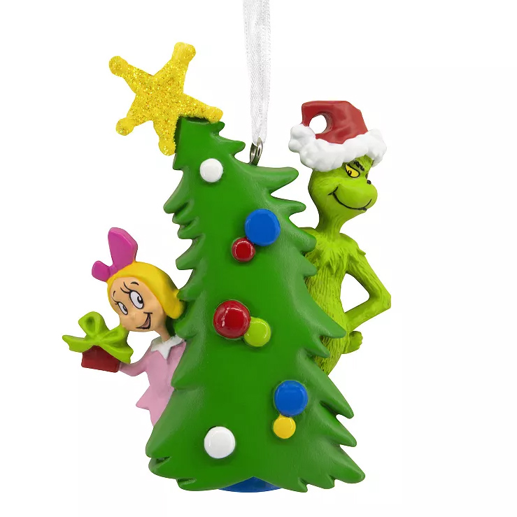 A colorful Christmas ornament featuring the Grinch in a Santa hat mischievously peeking around a decorated Christmas tree, with Cindy Lou Who cheerfully holding a gift and peering from the other side.