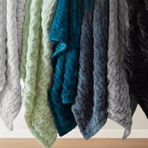 A selection of plush, textured blankets in various colors, including shades of gray, green, and blue, is displayed hanging in a row.