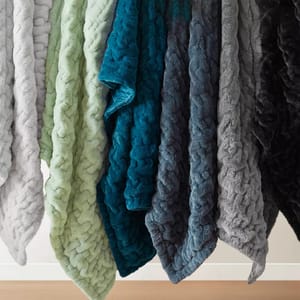 A selection of plush, textured blankets in various colors, including shades of gray, green, and blue, is displayed hanging in a row.